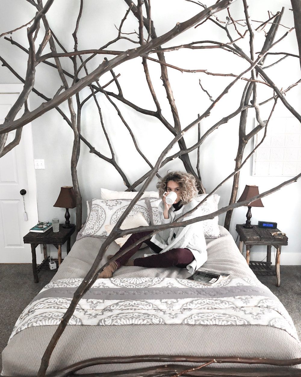 Woman drinking coffee while sitting on the bed with a book. Image is shot from the end of the bed through the tree branch bed frame.