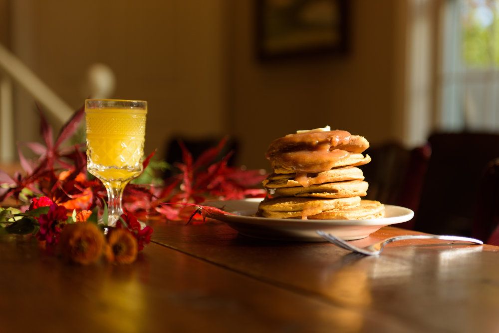 A tall stack of pancakes, covered in apples and syrup, sits next to a glass of fresh orange juice. In the background, fall leaves can be seen decorating the table.