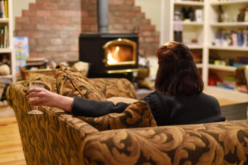 A woman is sitting on a comfortable couch in front of the wood-burning fireplace while drinking a glass of wine.