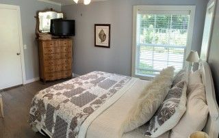 The primary bedroom shows a queen sized bed and large dresser with a tv. There are multiple windows that allow for natural light.