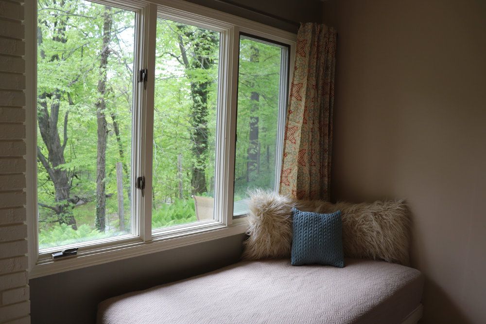 A day bed, decorated in pillows, sits next to three large windows.