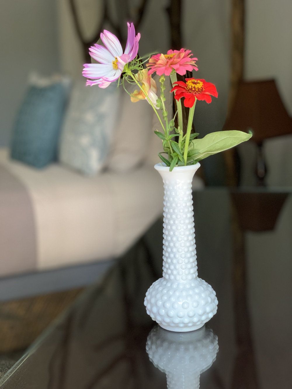 A multi-colored flower bouquet in a white vase sits on a desk. In the background, the bed and the nightstand are pictured.