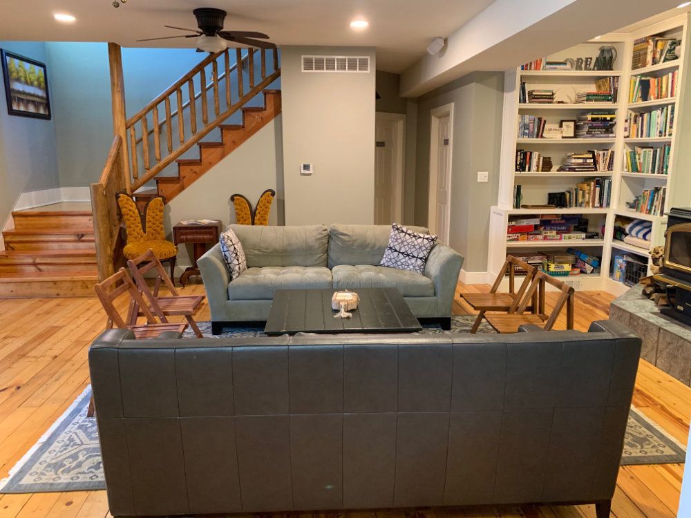 The main common room shows two couches, chairs, a coffee table, and bookshelves. In the background you can see the staircase leading up to the American Hornbeam and Sassafras rooms.