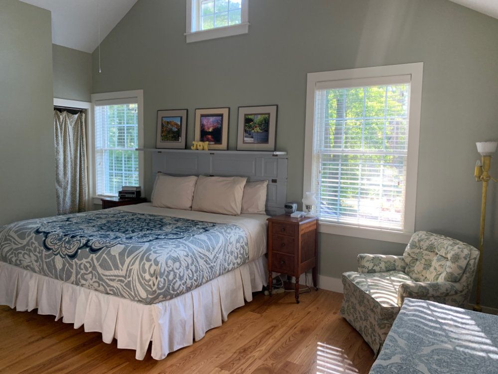 Second view of the primary bedroom. This image shows the king-sized bed, nightstands, and comfortable reading chair. The room is lit by three large windows and the walls are decorated with photos of the gardens.
