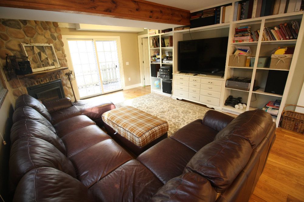 This image of the Inkeeper's living room shows 6+ person leather couch and ottoman, flatscreen tv, and bookshelves filled with games, books, and decor. To the left, a stone gas fireplace and large sliding doors can be seen.
