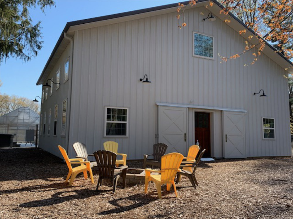 A view of The Barn's firepit in front of the building. Grey and yellow chairs surround the pit.