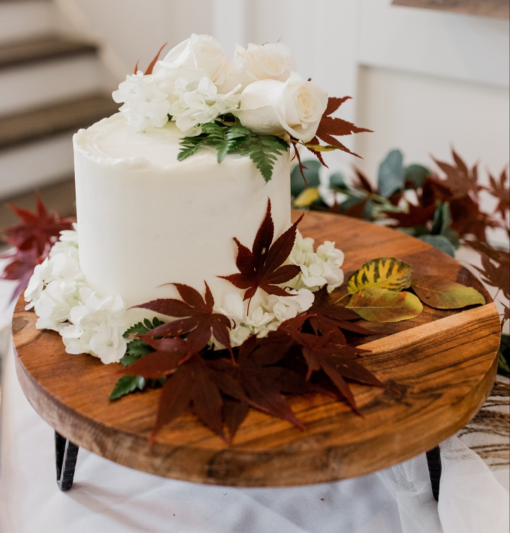 A white wedding cake decorated in fall leaves sits on a pedestal.