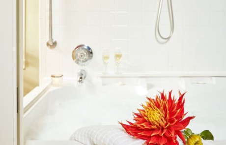 Large bubble bath with beautiful large red dahlia on a bath towel draped over the side of the jacuzzi. Wine glasses can be seen in the background.