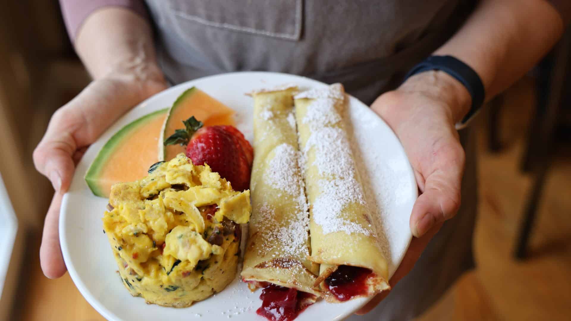 someone holding a white plate of breakfast. the plate has scrambled eggs and two beautifully rolled crepes