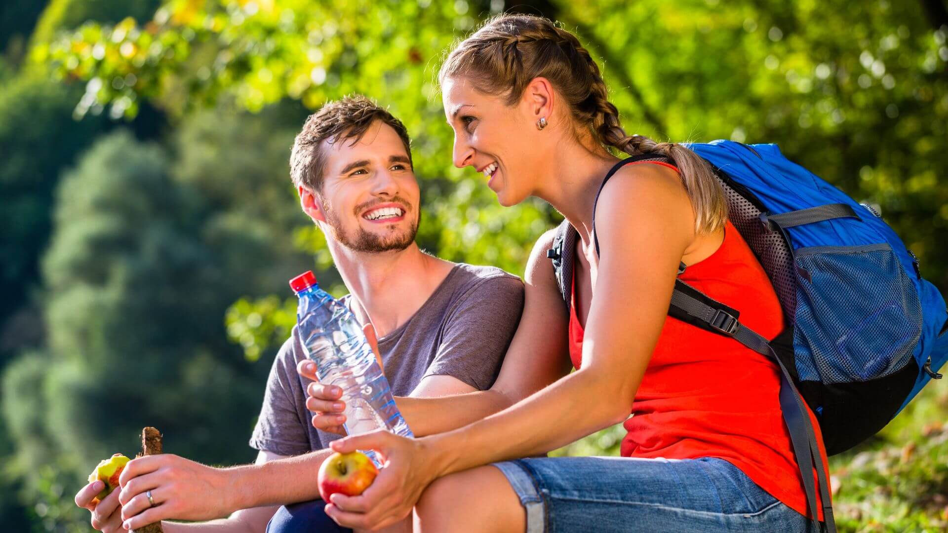 happy hiking couple sitting on a boulder talking and enjoying an apple and bottle of water