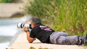 nature photographer lying prone on the sand with a camera and a large distance lens to his face