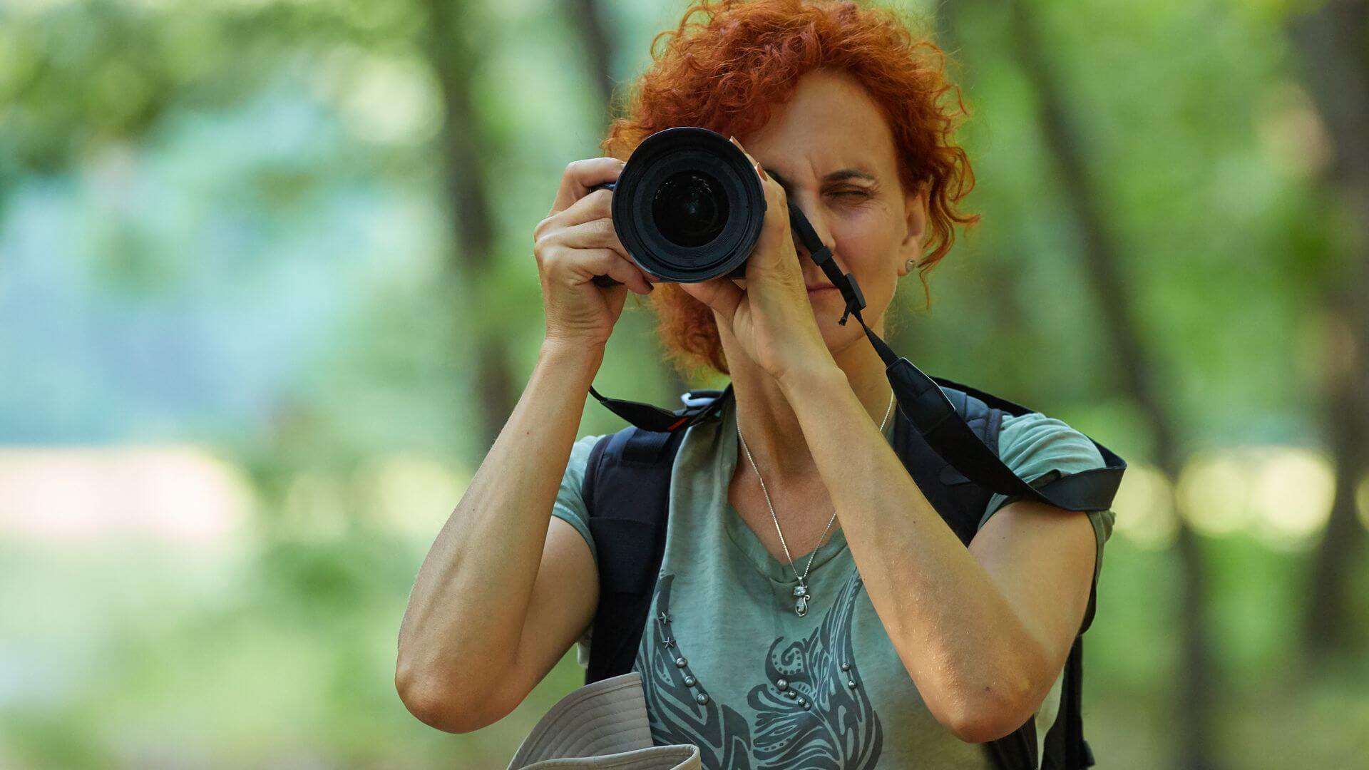 a woman looking through a camera and facing forward while standing in a blurred backdrop of nature