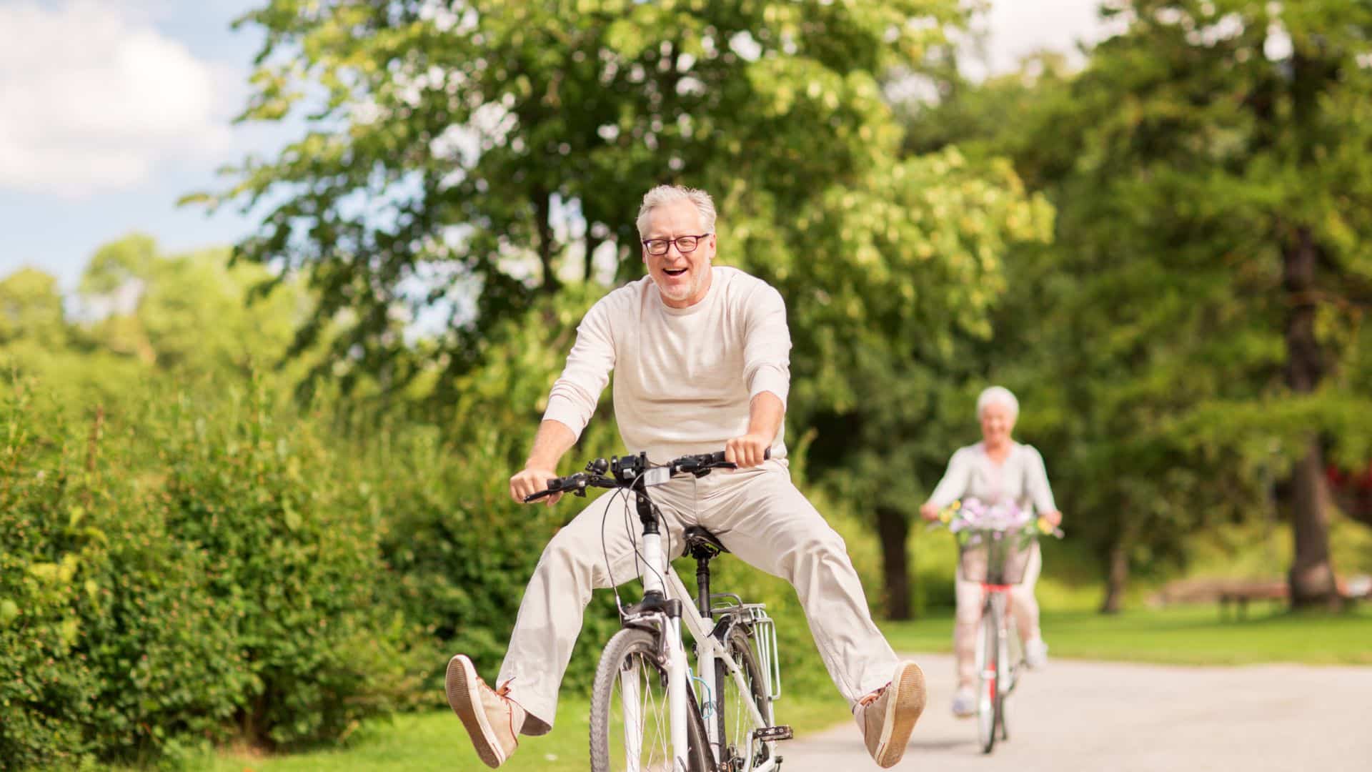 playful senior man smiles while riding bike through a park bike trail with legs off his pedals a blurred senior woman trails behind in the backdrop