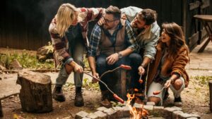group of laughing friends huddled near the fire pit and roasting hot dogs on sticks