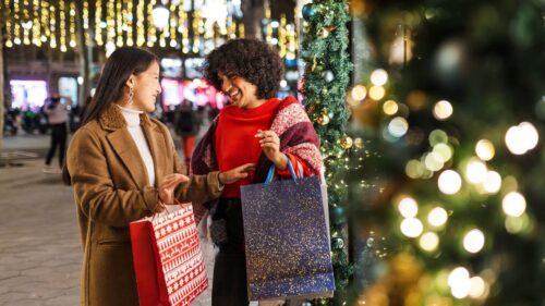 two diverse women standing outside and holding holiday shopping bags and talking while surrounded by sparkling lights and holiday decor