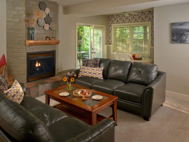 Living room with leather couches, coffee table, fireplace with wood mantle and table with four chairs by large windows