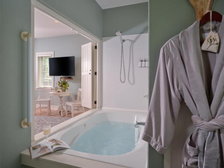 Bathtub with a shower head above it, a grey bathrobe hanging to the side, and a small white table and chairs and TV in another part of the room.