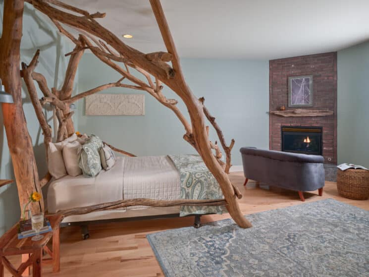 A bedroom with a bed with a unique driftwood frame, wood floors, a blue tapestry area rug, and a sitting area with a blue loveseat in front of a fireplace in a brick hearth.