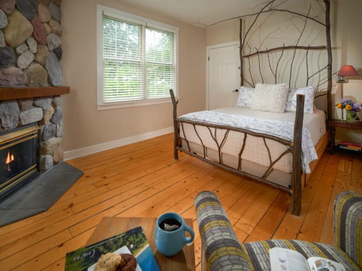 A bedroom with knotty pine floors, a bed framed with ironwood, a fireplace in a stone hearth, and a table and chair with a blue cup of tea and a muffin on the table.