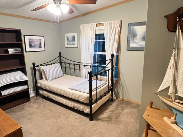 A bedroom with a wrought iron trundle bed and a bookshelf with extra bedding and an alarm clock.