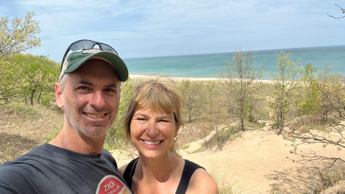 Smiling couple taking a selfie on a sandy dune with the turquoise sea in the background.