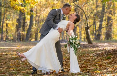 A groom dipping his bride outdoors surrounded by trees in fall colors