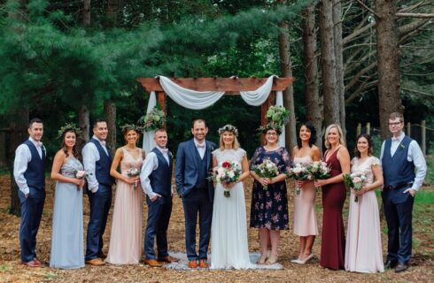 A wedding party gathered for a photo in the woods in front of a wooden arch