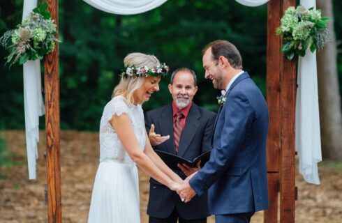 A bride and groom laughing together in front of an officiant, below a wooden arch in the woods