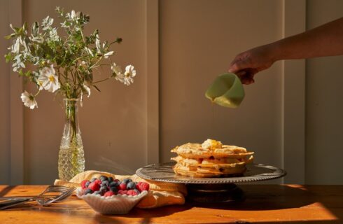 A person pouring syrup on a stack of waffles that are on a platter next to a bowl of berries and vase of flowers