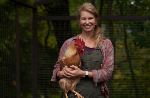 A wwoman in green overalls and a plaid shirt standing outside holding a rooster