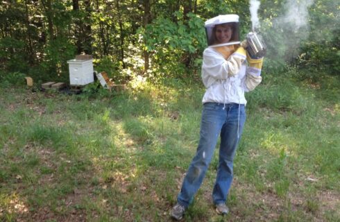 A woman in a bee suit holding a smoker in front of a hive in the woods