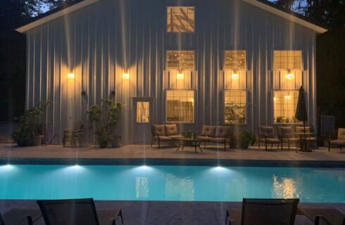 An outdoor pool at night next to a large white barn with golden light coming through the windows