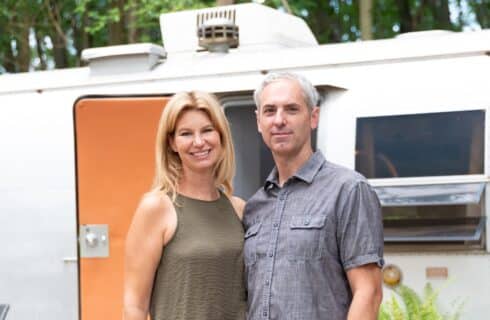 A man and woman standing outdoors in front of a vintage airstream camper
