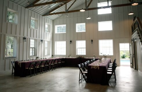 Large white barn with lots of window, wood beams, string lights and a U-shaped table set up for a meal