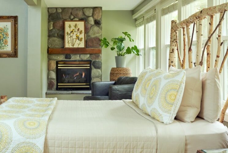 A bedroom full of light featuring a white birch bed with a long wall of windows, and leather chair by a stone fireplace