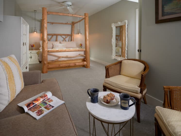 A bedroom with a knotty pine log 4 poster bed, a white wardrobe and mirror, and a sitting room with a loveseat, 2 armchairs, and a small table with a plate of muffins and 2 blue coffee mugs