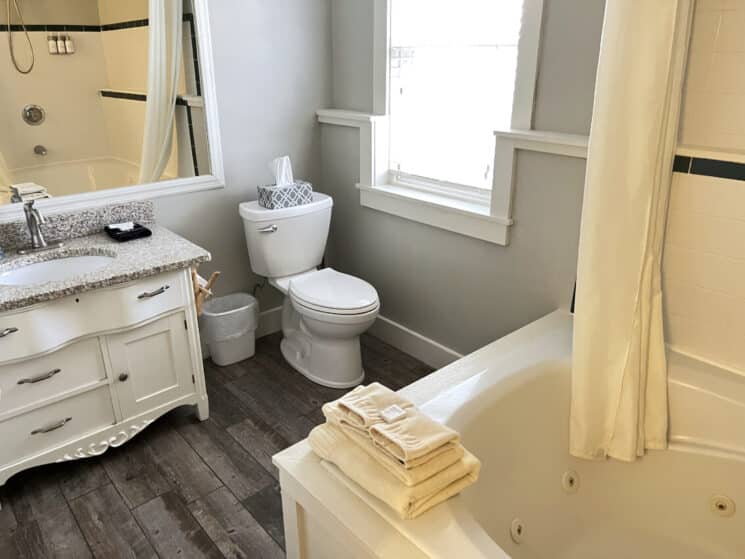 A bathroom with a white vanity with grey granite counter, wood floors, and a jetted bathtub with towels on the corner.