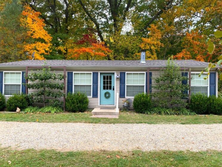 Front view of a ranch style cottageg with grey siding and navy blue shutters surrounded by trees
