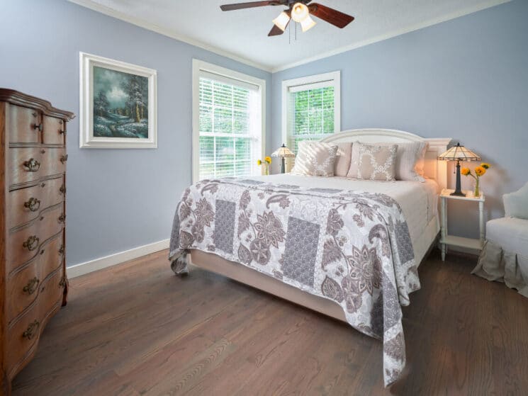 Bedroom in soft grey colors with queen bed, dresser with lamp and sitting chair by doorway to a bathroom