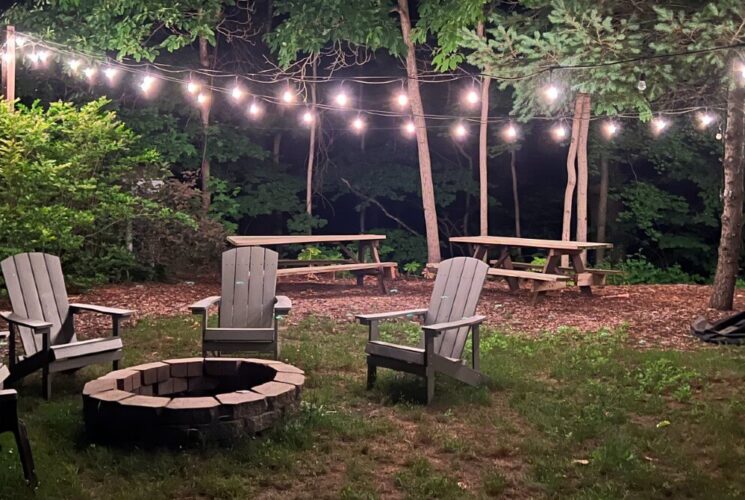 Outdoor firepit area with four Adirondack chairs, two picnic tables and string lights in the trees