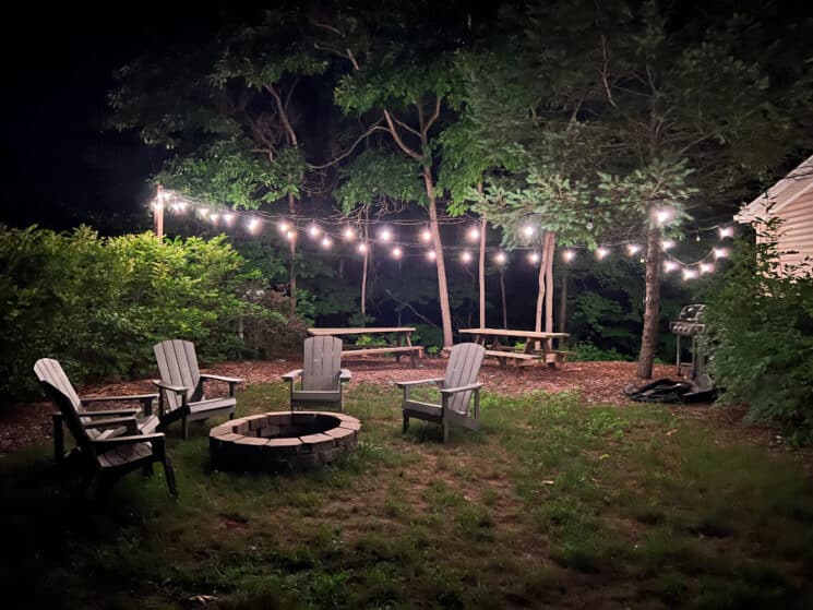 Outdoor firepit area with four Adirondack chairs, two picnic tables and string lights in the trees