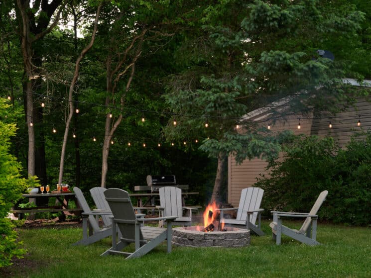 A grassy outdoor area with a firepit and grey adirondack chairs around them, a BBQ grill and picnic table in the background, with twinkle lights strung above them.