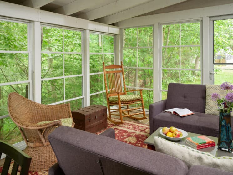 A screened in porch with brown sofas facing each other with a green coffee table in between them with a bowl of apples, some books and a blue vase with purple flowers, along with a wicker chair and wooden rocking chair, and a trunk in between them.
