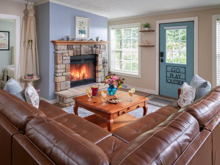 Living room with a brown leather couch, coffee table with a plate of cookies and glasses of iced tea, a fireplace with a fire going, and a blue door to outside that says GO PLAY OUTSIDE
