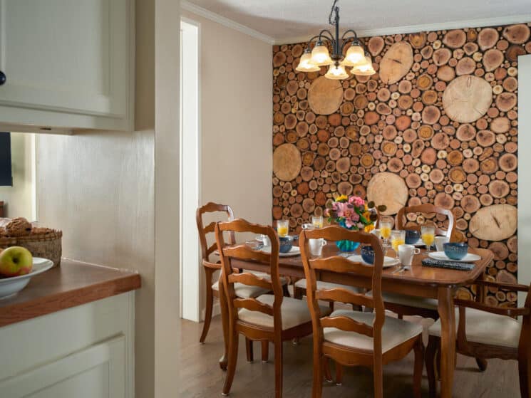 A wood dining table and chairs set for breakfast, log wallpaper, and a passthrough to another room with a bowl of fruit and basket of muffins on the counter.