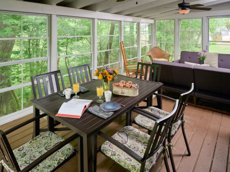 Outdoor screened in porch with black table and chairs with a basket of muffins, plates, glasses of orange juice and coffee cups, and a sitting area with 2 purple sofas facing each other, along with a rocking chair and wicker chair.