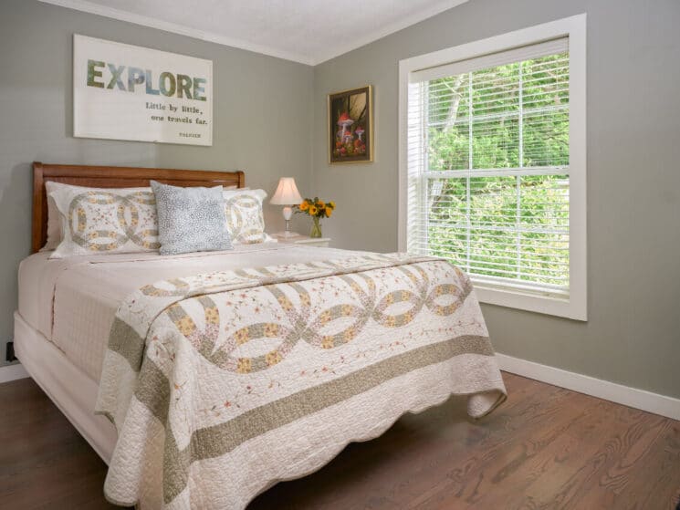 Bedroom with a bed with quilt, wood headboard, artwork and large window with blinds