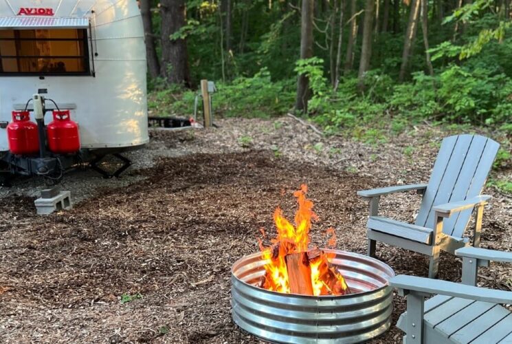 Vintage airstream camper in a wooded area with two Adirondack chairs by a roaring fire