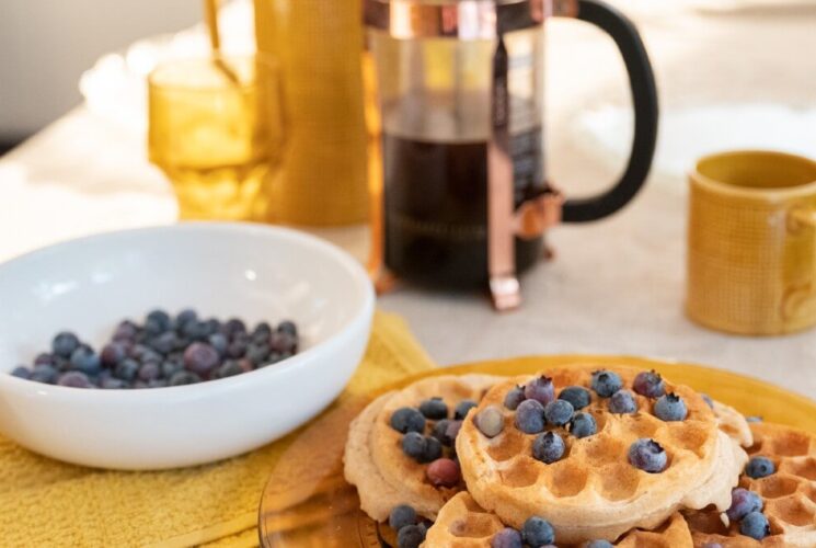 Plate of golden waffles with blueberries next to a french press of cofee and coffee mugs