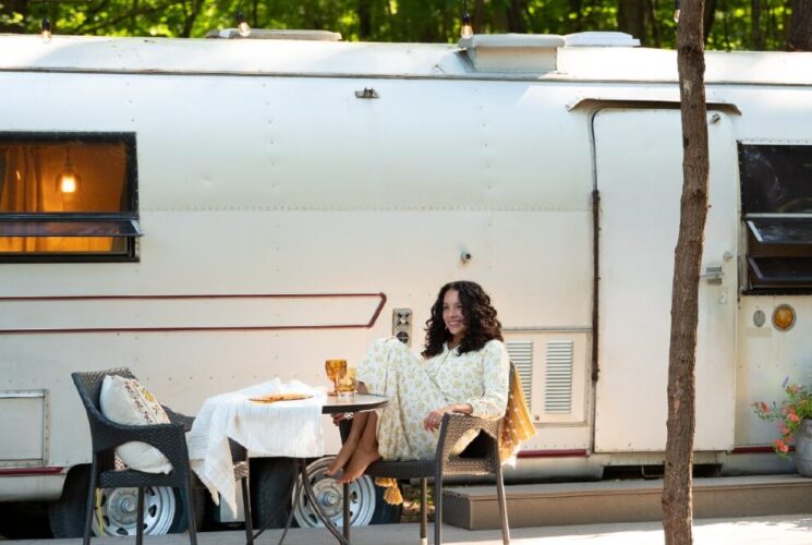 A woman sitting outdoors at a table in front of a large vintage airstream camper.